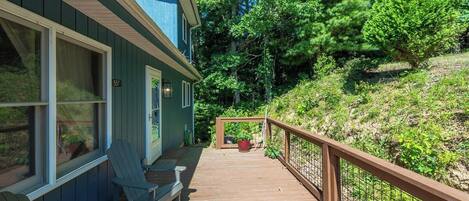 Une terrasse en bois mène à une maison à l’extérieur bleu, qui est entourée de verdure.