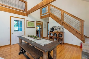 A wooden staircase with a glass railing, a dining table with a lace table runner, and a kitchen area in the background. - Cozy Cabin in town with a View! Hot Tub+Grill+WiFi (Burnsville)