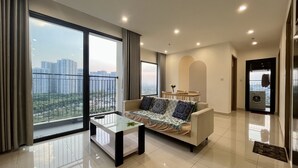 A modern living room with a sofa, a small table, and a view of high-rise buildings through large windows. - Yumy Homestay Grand Park City (Ho Chi Minh City)