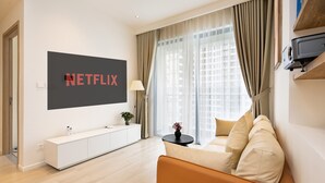 A modern living room with a large TV displaying "NETFLIX," a white console table, a sofa, and a window with sheer curtains. - Yumy Homestay Grand Park City (Ho Chi Minh City)