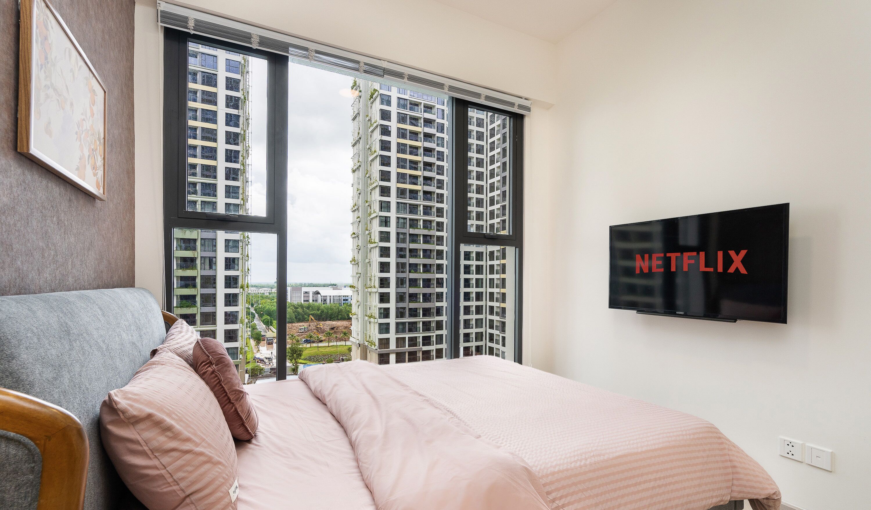 Habitación de hotel con cama, sofá, televisor montado en la pared y un ventanal con vistas a edificios altos.
