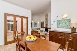 Un coin repas avec une table et des chaises en bois, un saladier rempli de fruits et une vue sur l’extérieur.