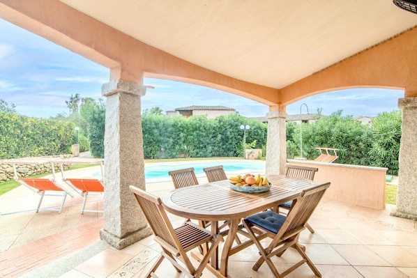 Une terrasse extérieure avec une table ronde en bois et des chaises, un saladier rempli de fruits, et une vue sur une piscine et la verdure environnante.
