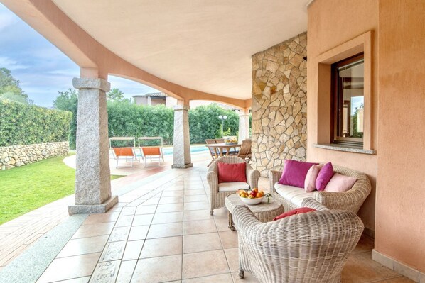 A covered patio area with wicker furniture, a stone wall, and a view of a swimming pool.