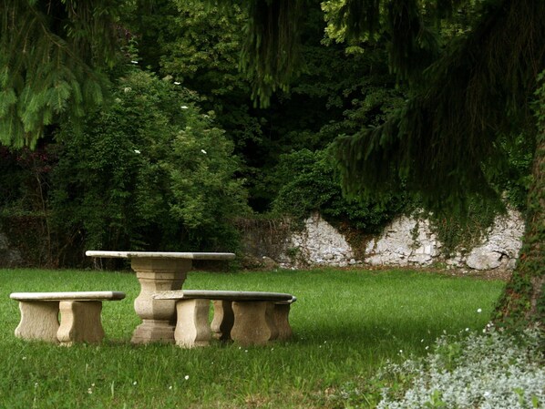 Une table et un banc en pierre, installés dans un jardin luxuriant, avec un feuillage dense et un mur de pierre en arrière-plan.