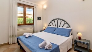 Een net opgemaakt bed met blauw-witte lakens, een klein nachtkastje met een lamp en een raam met gordijnen.