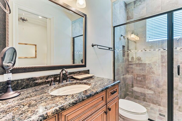 A bathroom with a granite countertop sink, a large mirror, a glass shower enclosure, and wooden cabinets.