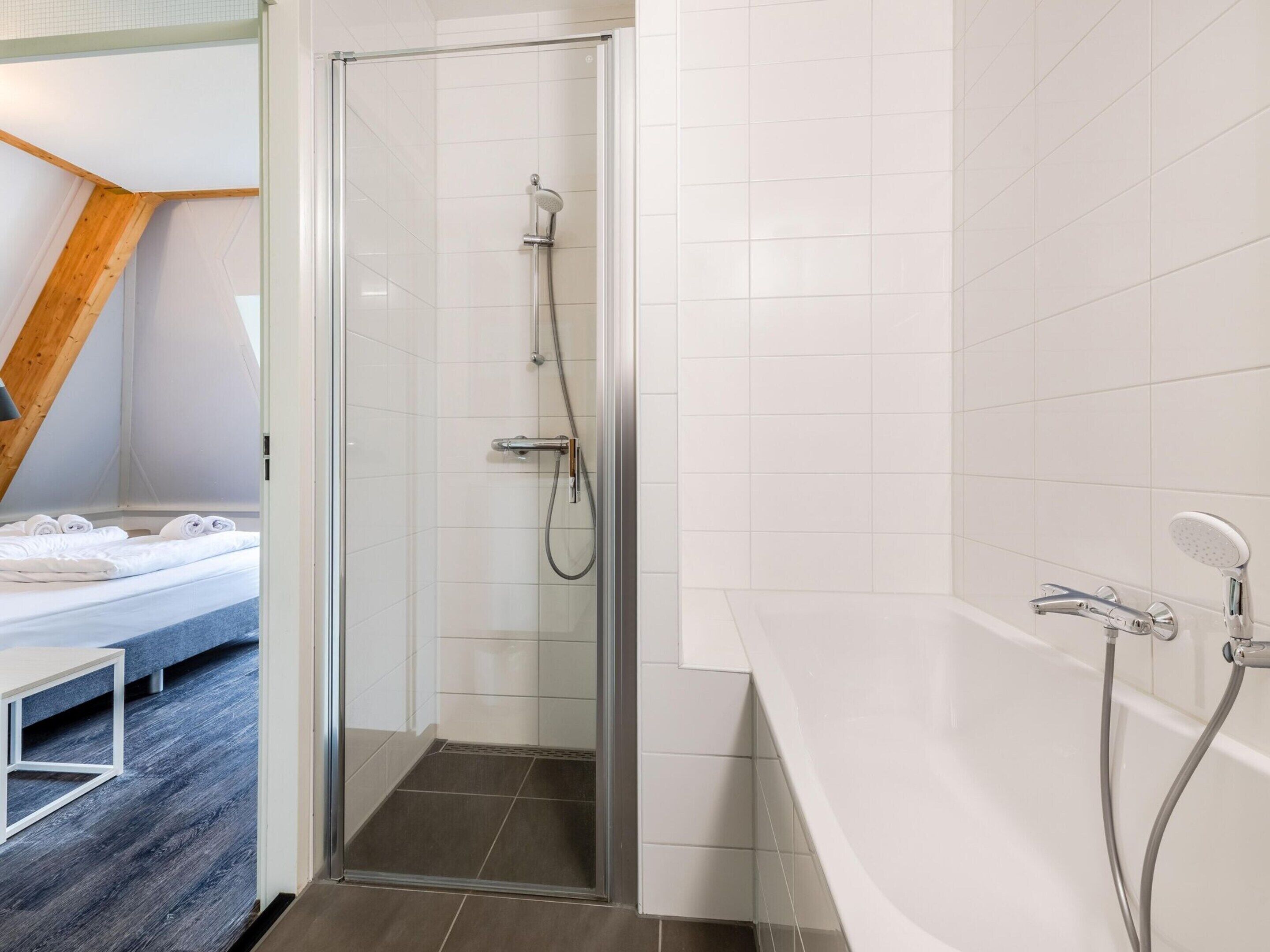 A modern bathroom with a glass shower enclosure, a bathtub with a handheld showerhead, and a bed with neatly made white linens in the adjacent room.
