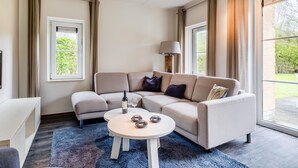 A modern living room with a beige sofa, a round coffee table, and a large window providing a view of greenery outside. - Villa in Banjaard Near Sandy North Sea Beach (Kamperland)