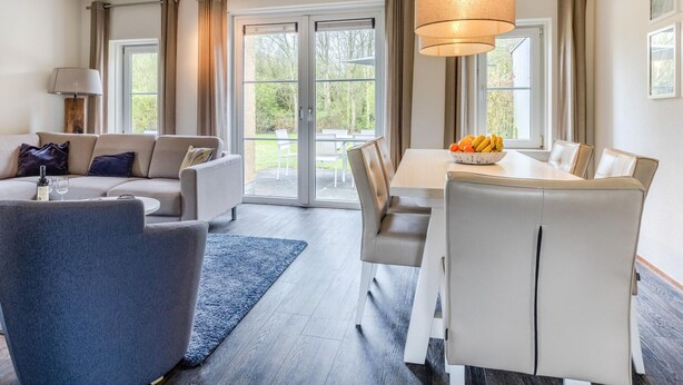 A modern living room with a sofa, a dining table with chairs, a blue rug, and a large window with curtains. - Villa in Banjaard Near Sandy North Sea Beach (Kamperland)