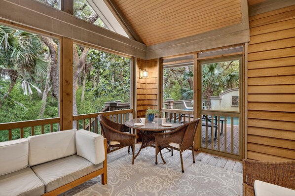 A covered outdoor seating area with a table and chairs, surrounded by wooden railings and large windows.