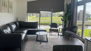 Una sala de estar moderna con un sofá de cuero negro, una mesa redonda de centro y dos sillones. Amplias ventanas ofrecen vistas a un patio con una fuente de agua y edificios.