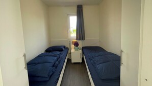 Una habitación pequeña y funcional con dos camas individuales, una mesita con un florero y una ventana con cortinas.