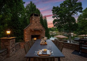 Un espace extérieur aménagé pour les repas, avec une cheminée en pierre, une table dressée pour deux personnes et une vue sur un lac et des arbres en arrière-plan.