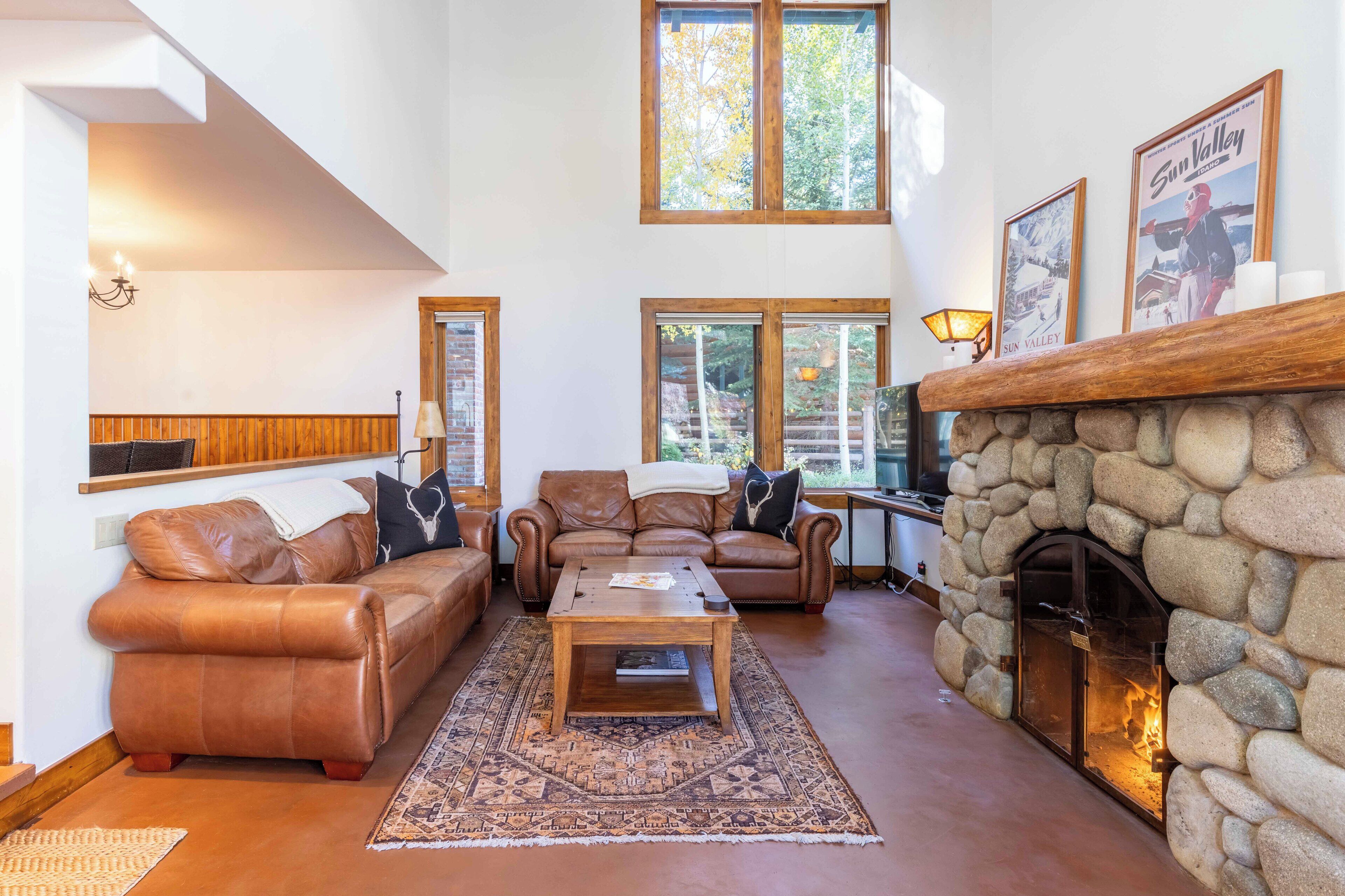 A cozy living room with a stone fireplace, leather sofas, and a large window overlooking a forest.