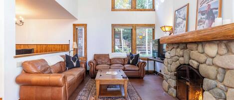 A cozy living room with a stone fireplace, leather sofas, and a large window overlooking a forest.