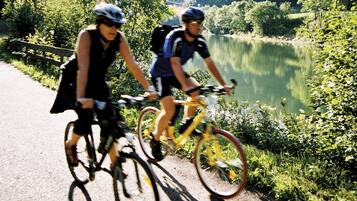 Two cyclists on a paved path beside a lake with trees and hills in the background.