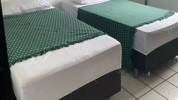 Quarto com duas camas, cada uma com colcha verde e almofadas brancas. Há uma janela com persiana de madeira e um ventilador fixado na parede.