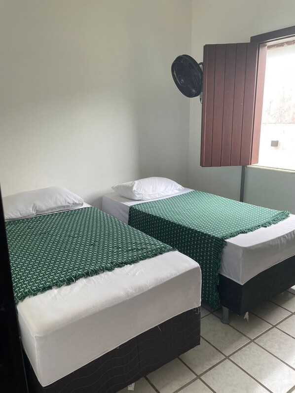 A room with two beds, each with a green bedspread and white pillows. There is a window with a wooden blind, and a wall-mounted fan. - Quarto com 2 camas de solteiros (Vera Cruz)