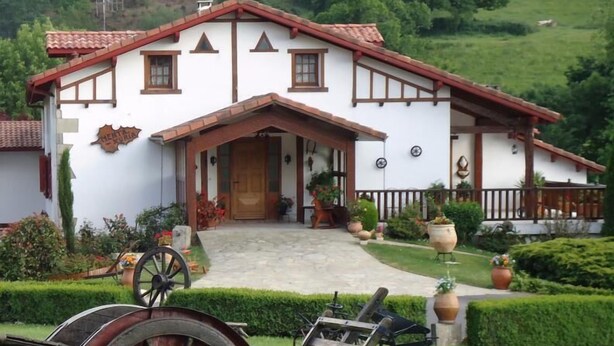 Ein traditionelles Haus mit weißer Fassade und rotem Dach, einer Holzveranda und einem Garten mit Topfpflanzen und einem Rasenwagen.