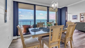 A dining area with a glass table and wooden chairs, large windows with a view of the sea, and a hanging light fixture. - Sailor's Delight 🌊 Deluxe 2-BR Condo @ Edgewater Beach & Golf Resort - Tower 1 (Panama City Beach)