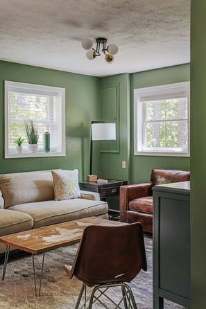 A living room with a green wall, two windows with white blinds, a beige sofa, a brown leather chair, a wooden coffee table, and a black cabinet.