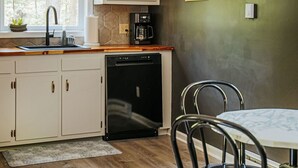 Une cuisine avec des meubles blancs, un lave-vaisselle, une machine à café, une fenêtre et un coin repas comprenant une table et des chaises.