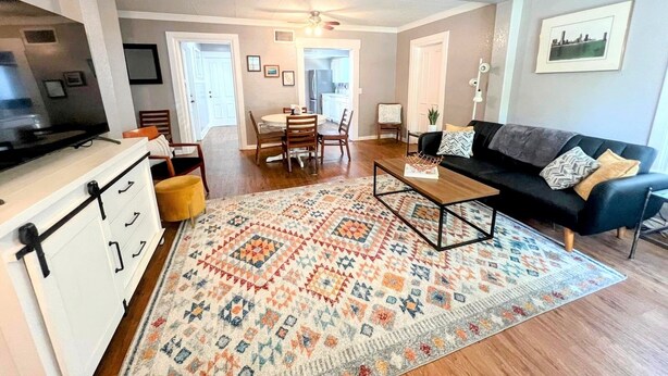 A spacious living room with a black sofa, wooden coffee table, and a colorful rug. There is a dining area with a table and chairs, and a kitchen in the background.