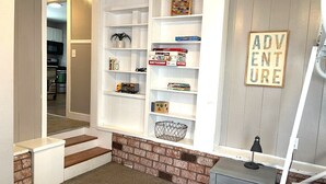 A home interior with a staircase leading to a kitchen, a bookshelf with various items, and a bed with a 'ADVENTURE' sign.