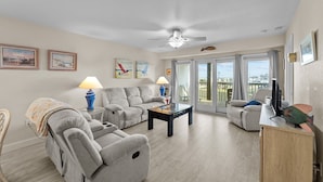 A spacious living room with a ceiling fan, a sofa, a coffee table, and a TV on a wooden cabinet. - 7081- Sea View - Rodanthe Condo | Coastal Beach Escape (Rodanthe)