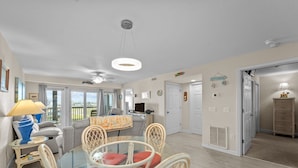 A spacious living area with a glass dining table, comfortable seating, a ceiling fan, and a view through large windows. - 7081- Sea View - Rodanthe Condo | Coastal Beach Escape (Rodanthe)