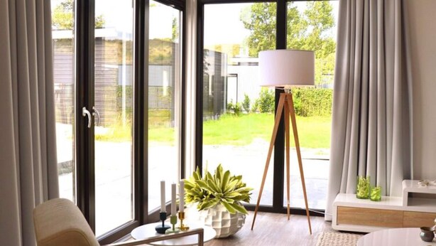 A modern living room with a large glass door leading to a garden, a round white table, a beige armchair, and a floor lamp. - Modern House With Dishwasher (Breskens)
