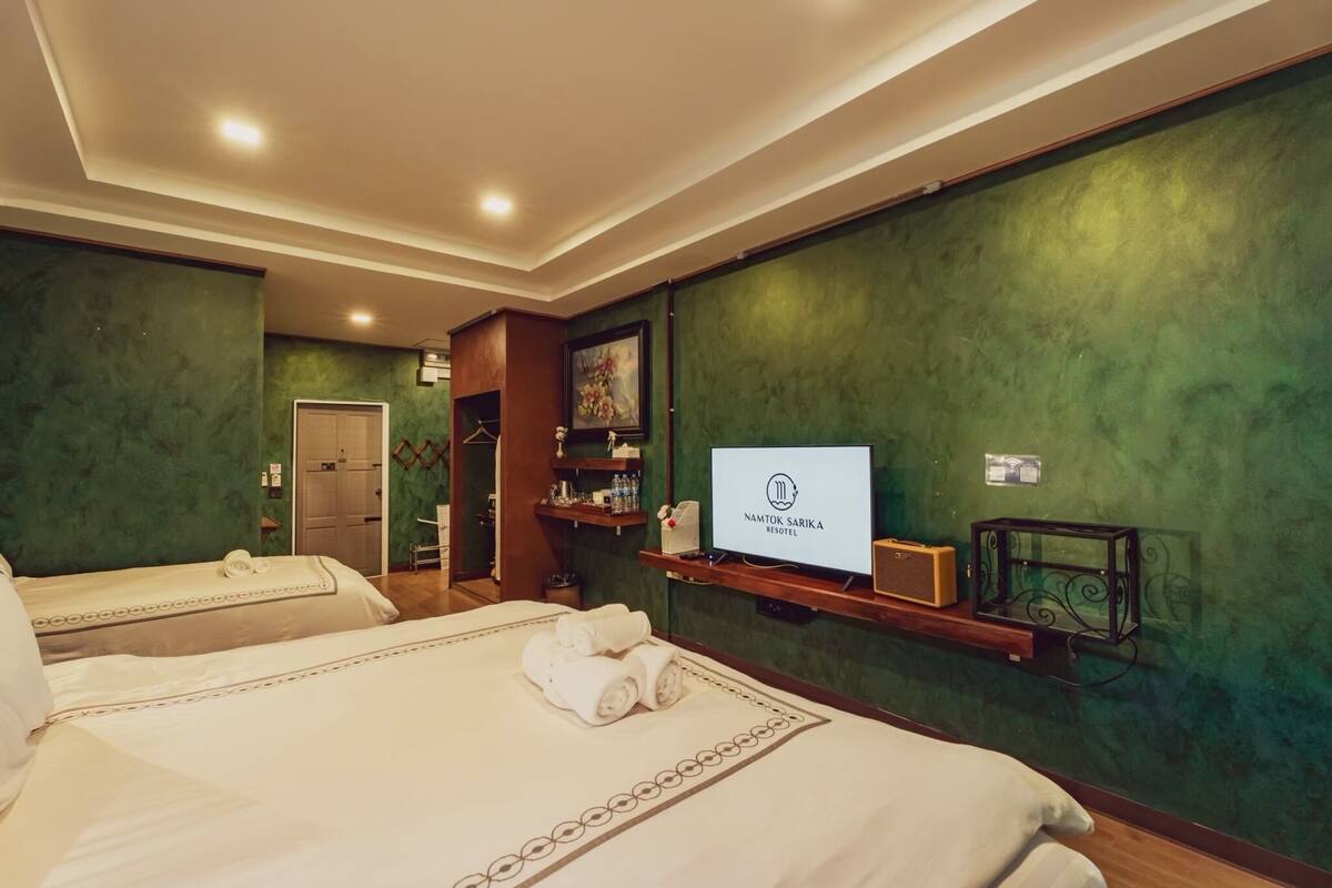Ein Hotelzimmer mit zwei Betten, einem Fernseher, einem Regal mit Pflegeprodukten und einer Wand mit grüner Struktur.