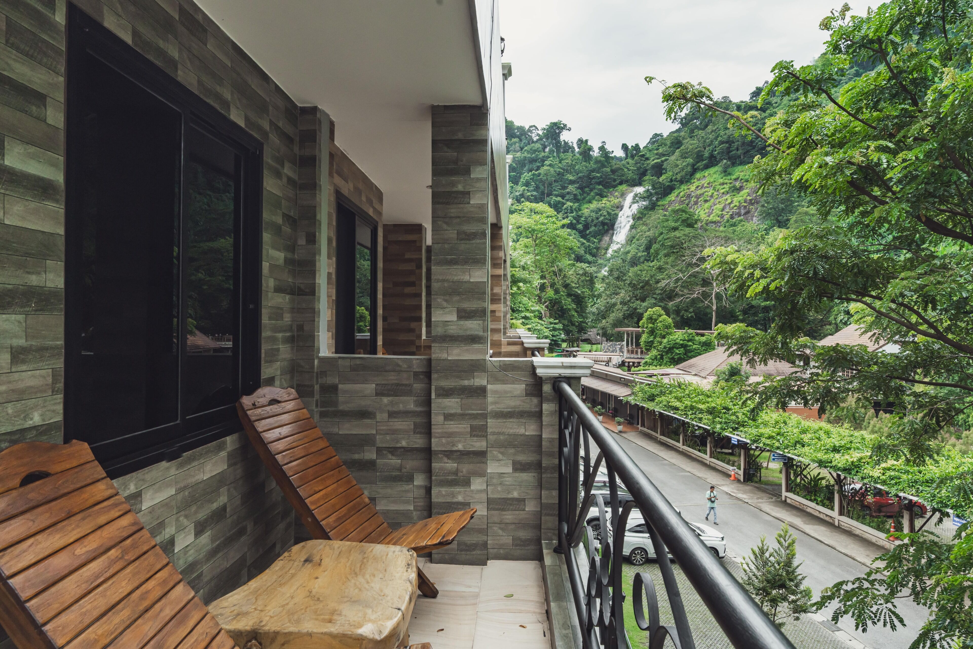 A balcony with wooden furniture, a view of a lush green landscape with a waterfall, and a street below.