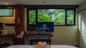 A hotel room with a bed, a TV displaying 'NAMTOK SARIKA RESOTEL,' a window with a view of a waterfall, and a desk with a microwave and a plant. - Namtok Sarika Resotel (Nakhon Nayok)