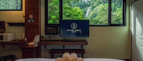 Habitación de hotel con cama, un televisor mostrando 'NAMTOK SARIKA RESOTEL', ventana con vistas a una cascada y escritorio con microondas y planta.