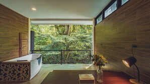 A modern bathroom with a bathtub, a wooden vanity, a glass vase with flowers, and a view of a forest through large windows. - Namtok Sarika Resotel (Nakhon Nayok)