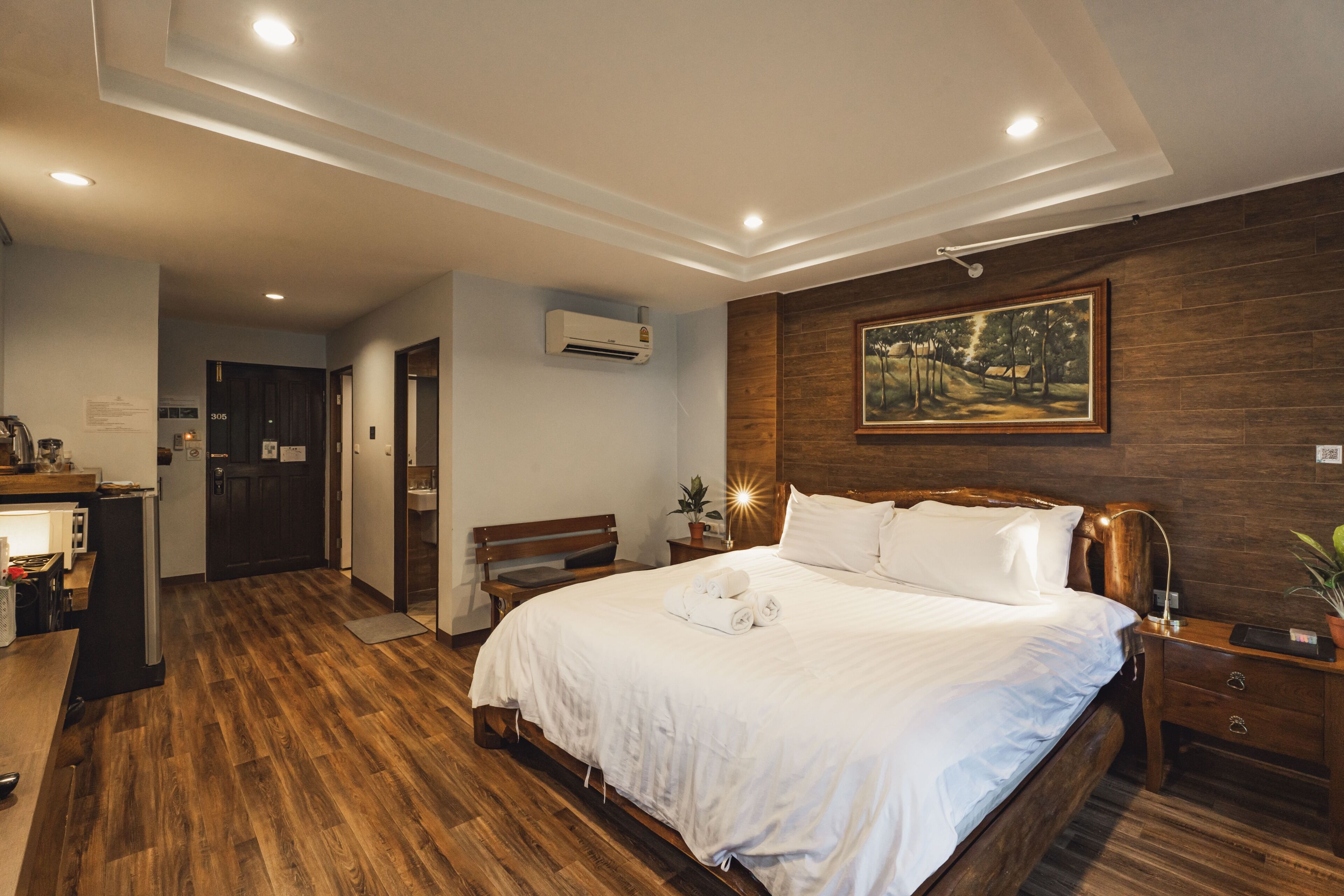 Habitación de hotel con suelo de madera, una cama grande con ropa blanca, una mesita de noche con lámpara, un banco y un cuadro en la pared.