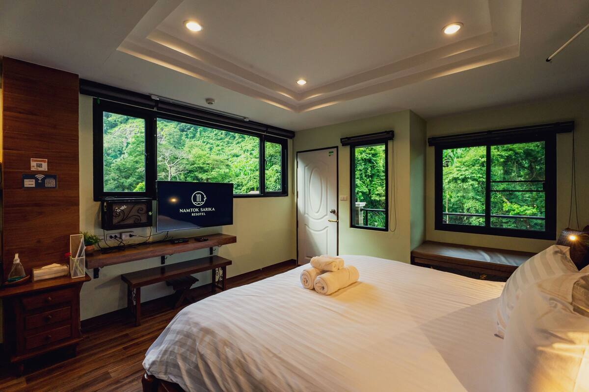 Ein Hotelzimmer mit Bett, Fernseher auf einem Holzschrank und großen Fenstern mit Blick auf einen Wald.