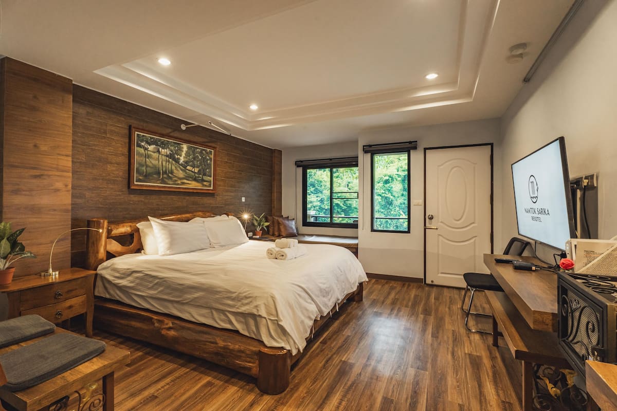 Ein Schlafzimmer mit einem hölzernen Bett, einem Schreibtisch, einem Computer und einem Fenster mit Blick auf Grünflächen.