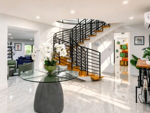 A modern interior with a glass table, a green vase with white flowers, a wooden staircase, and a seating area with a blue sofa. - Villa Palma: Heart of Miami | 5 min to Miracle Mile (Miami)