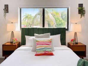 A hotel room with a large bed, two nightstands, a window with a view of palm trees, and two wall-mounted lamps. - Villa Palma: Heart of Miami | 5 min to Miracle Mile (Miami)