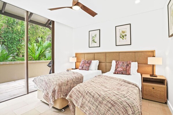 A hotel room with two beds, a ceiling fan, a balcony with a view of greenery, and framed artwork on the walls.
