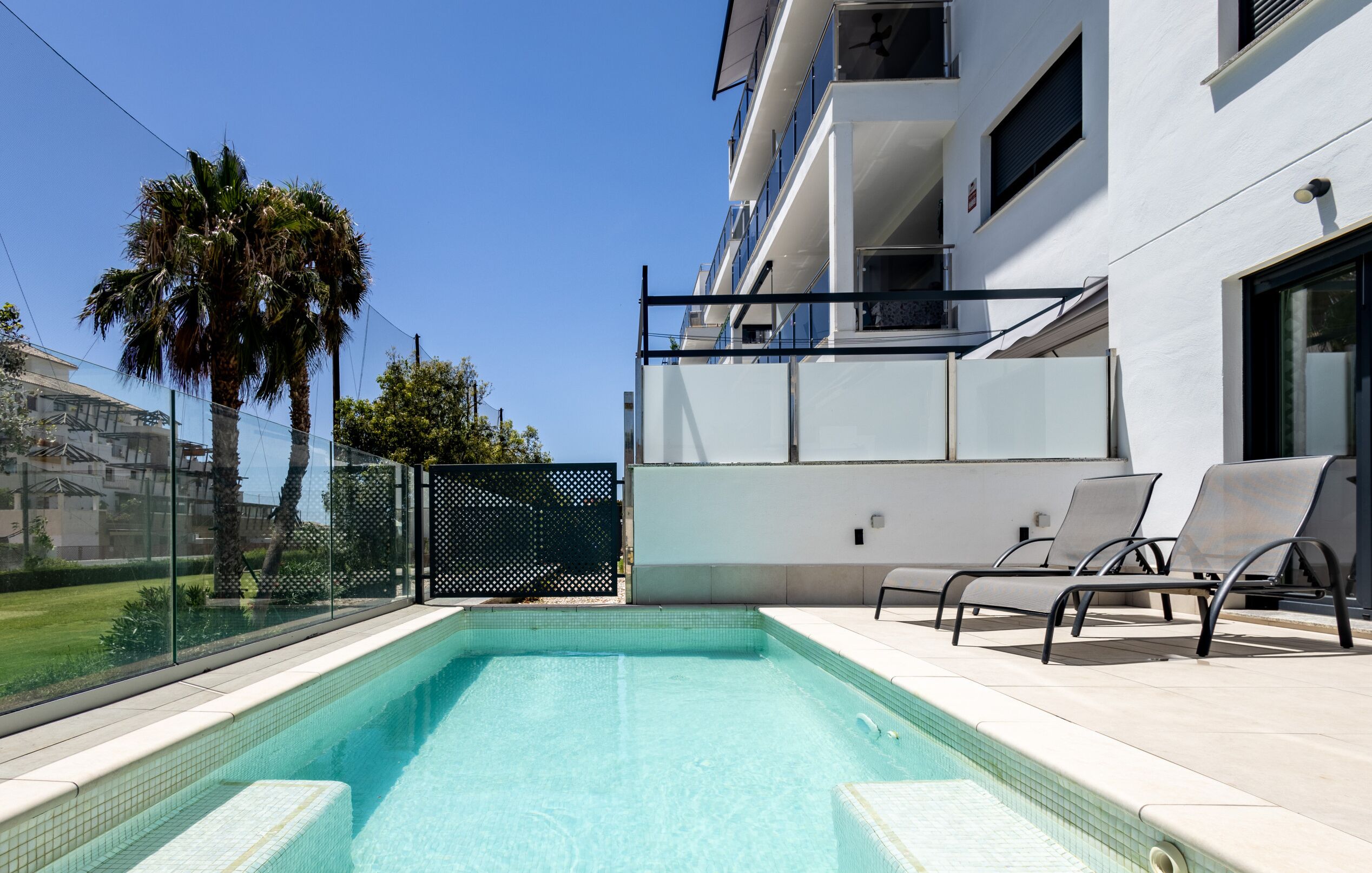 Una piscina exterior moderna amb aigua blava cristal·lina, envoltada d'un edifici blanc amb balcons i una tanca de vidre.