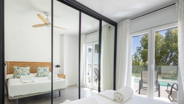 Ein modernes Schlafzimmer mit Bett, Deckenventilator und Blick auf einen Swimmingpool durch große Glasüren.