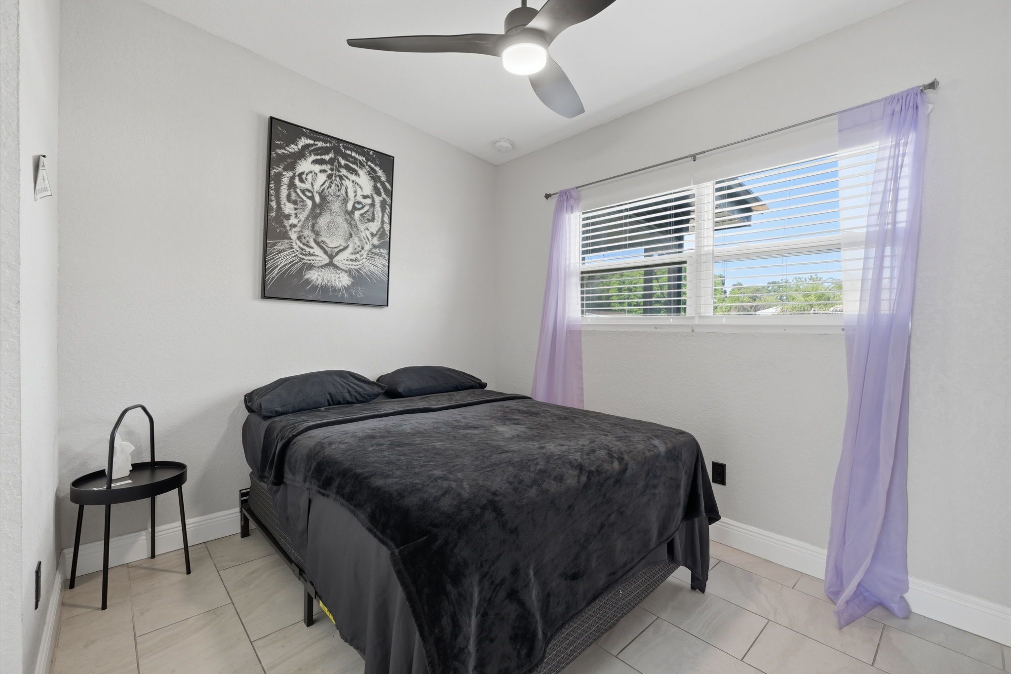 Une chambre à coucher avec un lit, un ventilateur de plafond, une fenêtre avec des stores et un tableau accroché au mur.