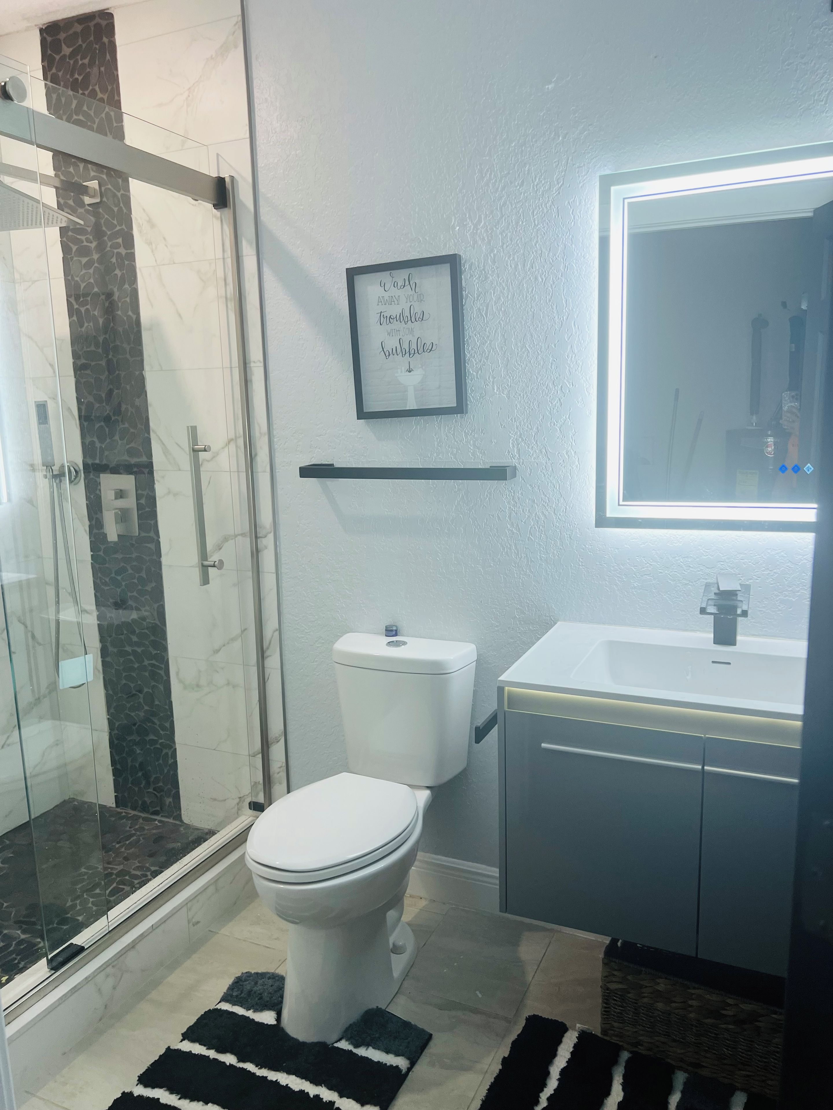 Ein Badezimmer mit Duschkabine aus Glas, Toilette, Waschbecken mit Spiegel und einem gerahmten Motivationsspruch an der Wand.