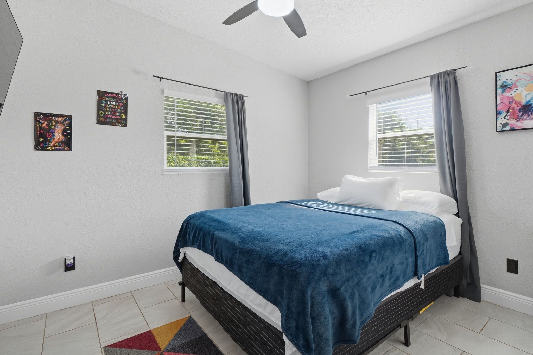 A bedroom with a bed, two windows with curtains, a ceiling fan, and a wall with two framed items.