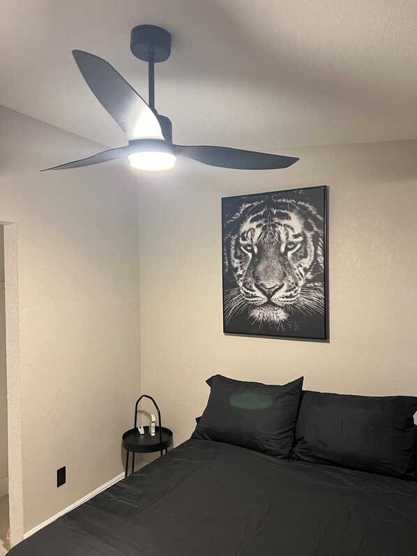 A bedroom with a bed, a ceiling fan, a wall-mounted picture, and a small bedside table.