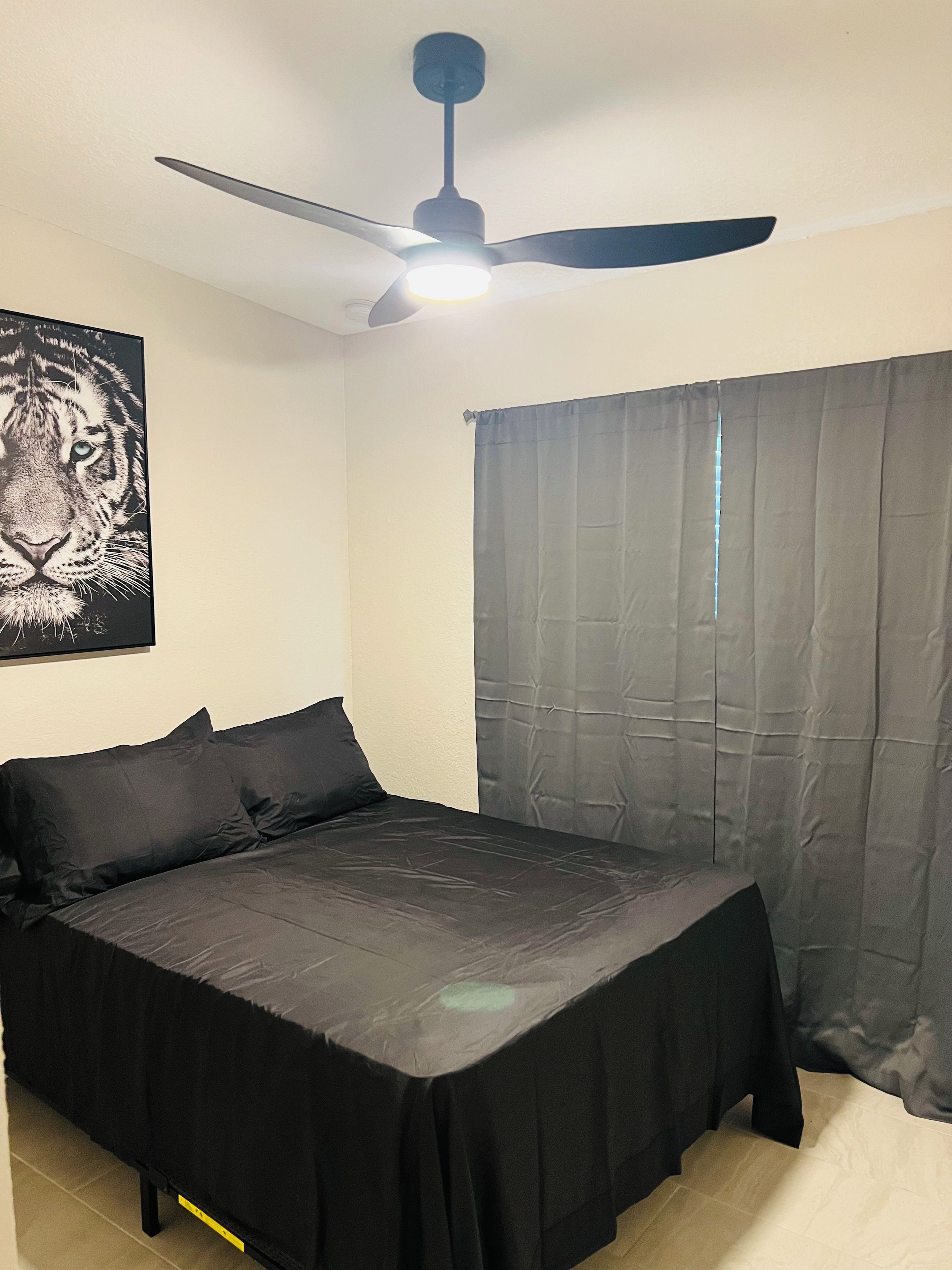 Une chambre à coucher avec un lit, un ventilateur de plafond, une fenêtre avec des rideaux et une affiche de tigre accrochée au mur.